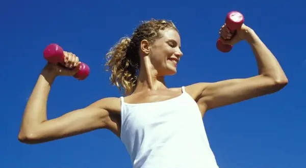 Femme souriante avec des haltères roses, montrant ses muscle bras. Elle porte un débardeur blanc sous un ciel bleu clair.