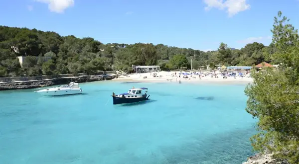 En solig dag på mallis Mallorca. Turkosblått vatten, en vit yacht och en blåvit båt i en vik med en livlig strand.