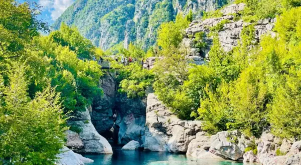 Aguas turquesas fluyen entre rocas y vegetación exuberante, bajo imponentes montañas en Albania. Un puente peatonal cruza el cañón, invitando a explorar las **montañas malditas de Albania**.