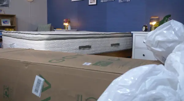 A mattress that expands when opened is shown, partially covered by a cardboard box and plastic packaging.