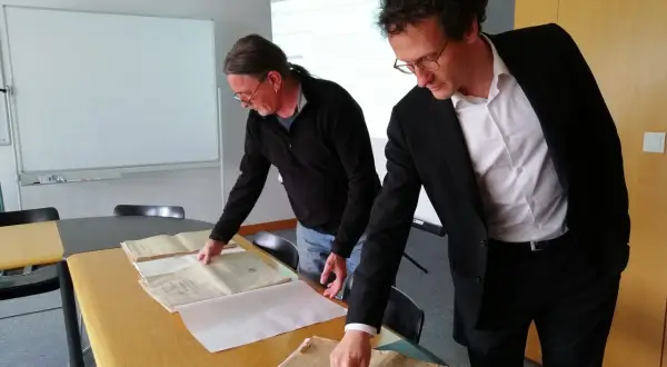 Deux hommes consultent des registres d'état civil aux archives départementales du Morbihan. L'un pointe un document ancien.