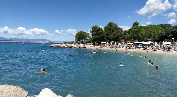 Crikvenica atrakcje: plaża pełna ludzi kąpiących się i opalających, błękitne morze, zielone drzewa i góry w tle.