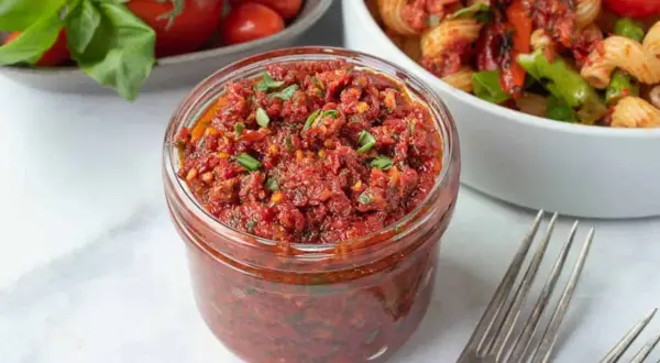 Vaso di pesto di pomodori secchi pugliese, pronto per condire la pasta. Freschi pomodori e basilico sullo sfondo.