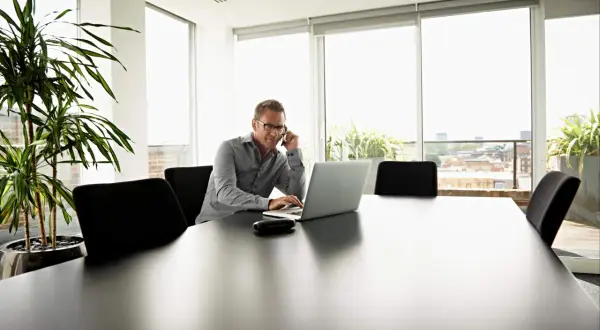 Biznesmen rozmawia przez telefon, pracując na laptopie. W tle okna z widokiem na miasto, a obok rośliny. To jego koszt uzyskania przychodu.