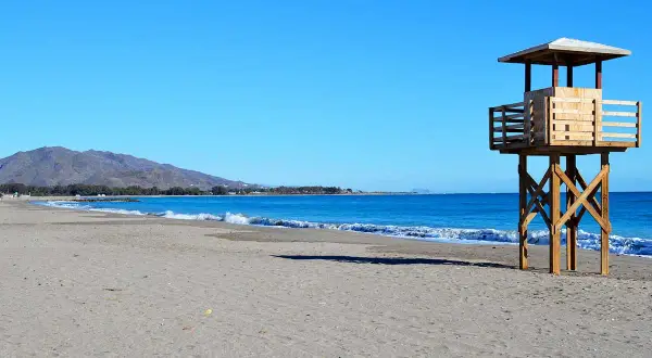 Torre de vigilancia de madera en la playa de Vera, con arena y mar azul. La playa de Vera tiene piedras, pero hoy el sol brilla.