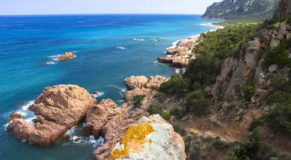 Scogliere rosa e mare turchese sulla costa ovest Sardegna. Un sentiero si snoda tra rocce e macchia mediterranea verso una spiaggia isolata.