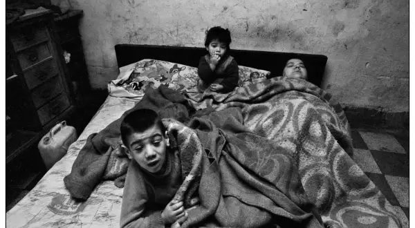 Bambini in un letto, un piccolo è seduto con le dita in bocca, un altro è sdraiato. Un'immagine toccante, una delle **Letizia Battaglia foto famose**.
