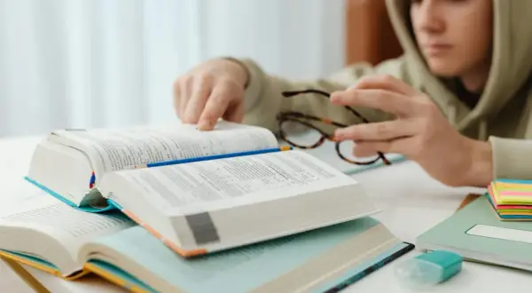 Chłopak w bluzie pochyla się nad książkami, analizując **motywy literackie**. Na stole leżą otwarte tomy, okulary i kolorowe karteczki.
