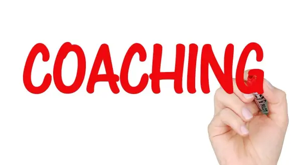 Ręka pisze czerwonym markerem słowo "COACHING". To proces rozwoju, który może obejmować lingwistykę.