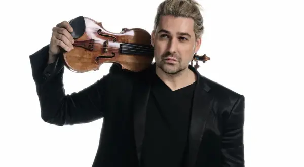David Garrett z blond włosami spiętymi w kucyk, w czarnym garniturze, trzymający skrzypce na ramieniu.