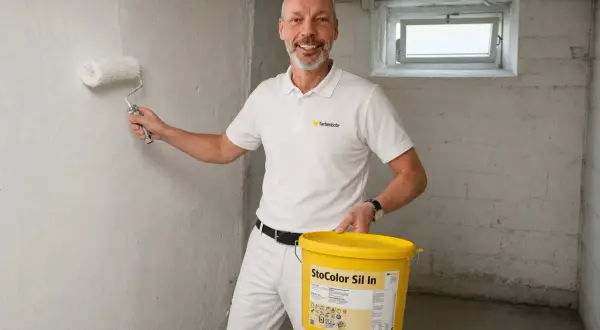 Mann streicht Kellerwand im Altbau mit weißer Farbe. Er hält eine gelbe Farbeimer-Marke "StoColor Sil In".