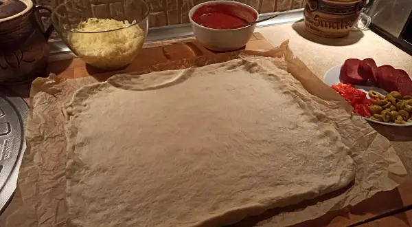 Gotowe ciasto na pizzę czeka na dodatki: ser, sos, salami, papryka i oliwki.