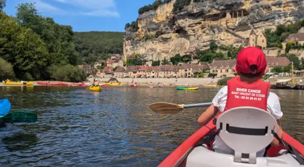 Enfant en canoë rouge, gilet de sauvetage "River Canoe", pour une visite insolite autour de Sarlat, avec des falaises et des maisons troglodytes en arrière-plan.