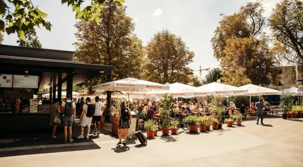 Sonniger Biergarten im Schlossgarten mit vielen Gästen unter schattenspendenden Schirmen.