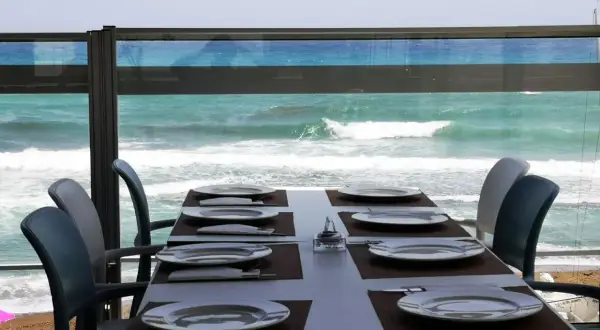 Mesa puesta en el chiringuito la sal, con platos y cubiertos, frente a un mar turquesa y olas rompiendo.