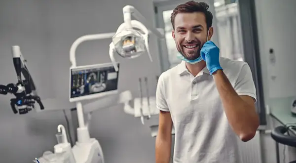 Uśmiechnięty dentysta w gabinecie, gotowy do pracy. Zastanawiasz się, ile trwa plombowanie zęba? To zależy od przypadku, ale zazwyczaj krótko!