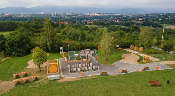 Nowoczesny plac zabaw i siłownia plenerowa, otoczone zielenią, z widokiem na miasto. W oddali widać trzy lipki.