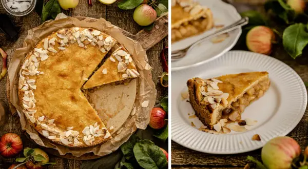 Ein goldbrauner gedeckter Apfelkuchen, bestreut mit Mandelblättchen, wird auf einem Holztisch präsentiert. Daneben liegen frische Äpfel und Blätter.