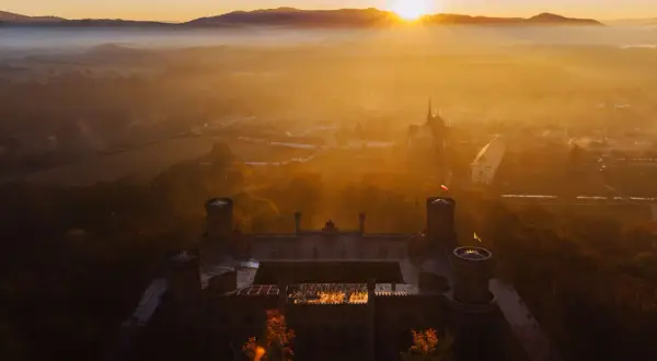 Zamek otoczony mgłą, skąpany w ciepłych promieniach **golden hour**. Słońce wschodzi nad wzgórzami, malując niebo na złoto.