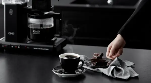 A black coffee maker brews coffee into a cup next to chocolate pieces and a napkin.
