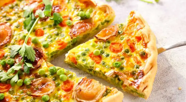 Un delicioso quiche de verduras, con guisantes, zanahoria y trozos de queso dorado, recién cortado y listo para servir.