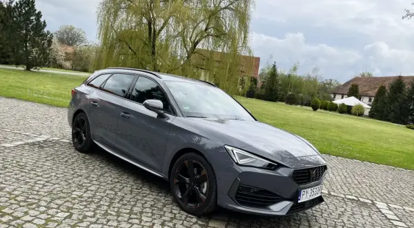 Szary Cupra kombi na brukowanym podjeździe, z zielonym trawnikiem i drzewami w tle.