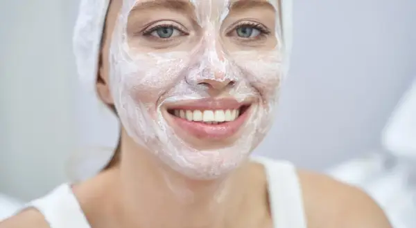 Lächelnde Frau mit weißer Gesichtsmaske und Handtuch auf dem Kopf. Wie oft Maske Gesicht? Regelmäßige Pflege für strahlende Haut.