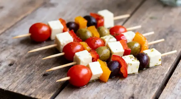 Des brochettes apéro colorées avec des tomates cerises, des cubes de fromage, des poivrons et des olives sur une table en bois.