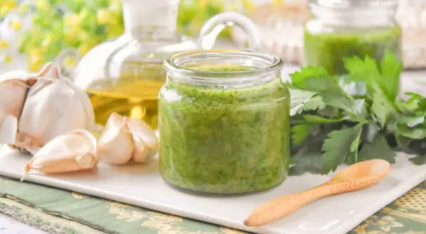 Un frasco de salsa mery verde vibrante, con ajo, aceite de oliva y perejil fresco alrededor.