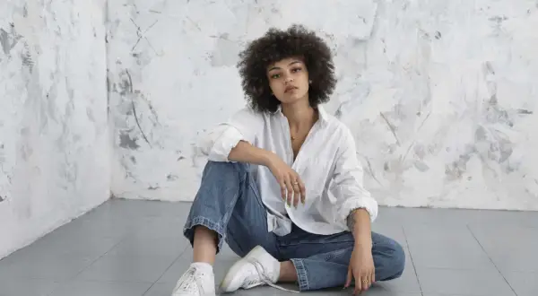 Kobieta z kręconymi włosami w białej koszuli i jeansach, siedząca na podłodze. Jej styl to klasyczny, basic look.