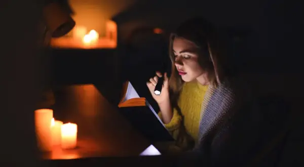 Dziewczyna czyta książkę przy świecach i latarce podczas blackoutu. Co to blackout? To nagłe przerwy w dostawie prądu.
