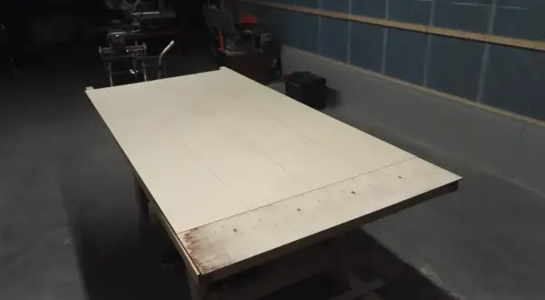 Stor skiva av plywood på ett arbetsbord, redo att bygga slagport. Verktyg syns i bakgrunden.