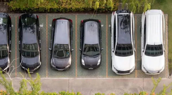 Sześć samochodów na parkingu. Odległość miejsca postojowego od okien domu jednorodzinnego jest niewielka, co widać po bliskości zieleni.