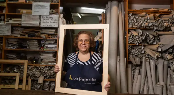 Mujer sonriente en taller, rodeada de rollos de tela y estanterías con materiales. Sostiene un marco de madera en rollo.