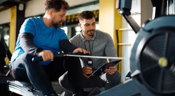 Dwóch mężczyzn na siłowni, jeden ćwiczy na wioślarzu, drugi z notatnikiem. Rozmawiają o tym, ile zarabia instruktor fitness.