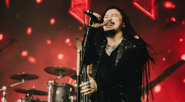 Wokalista zespołu rockowego na scenie, z mikrofonem w dłoni, w tle czerwone światła i perkusja. Koncert w Hali Stulecia we Wrocławiu.