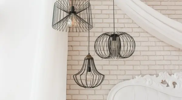 Nowoczesne **lampa pająk aranżacje** w loftowym stylu. Trzy wiszące lampy z drucianymi kloszami na tle ceglanej ściany i białej zasłony.