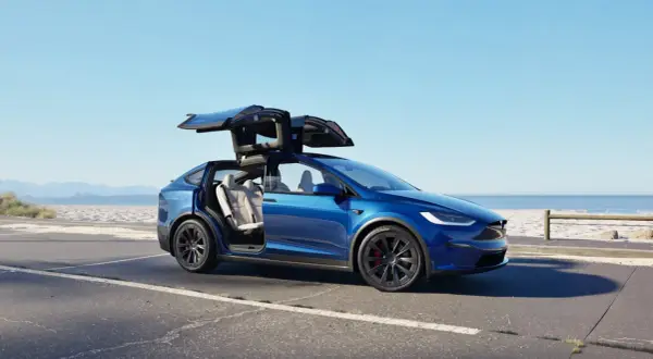 Niebieski Tesla Model X z otwartymi drzwiami typu falcon wing na tle plaży. Sprawdź aktualną tesla model x cena.