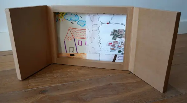 Un kamishibai casero muestra dibujos infantiles: una casa, un paisaje rural y un pueblo con un tren.