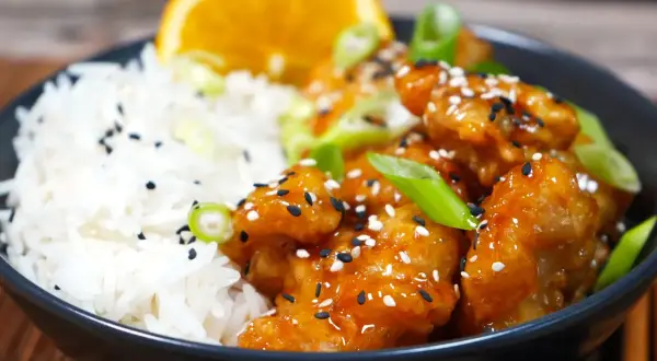 En skål med ris och krispig, glaserad orange chicken, toppad med sesamfrön och salladslök. En apelsinskiva vilar bakom.