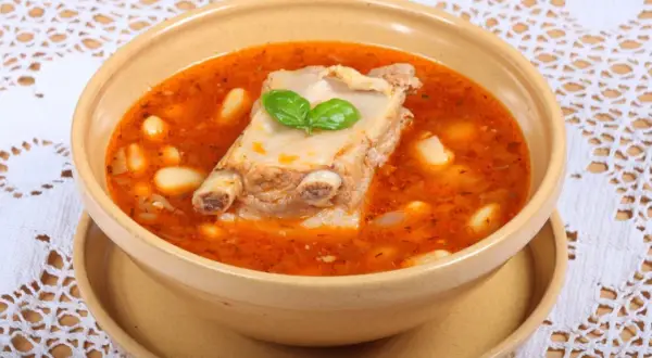 Aromaticzna zupa na żeberkach z fasolą i ziołami, podana w glinianej misce na ozdobnym obrusie.