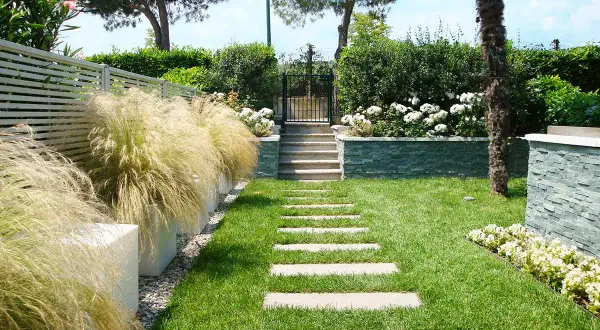 Un bellissimo giardino progetto con un sentiero di lastre di pietra, erba verde, cespugli fioriti e un muretto in pietra.