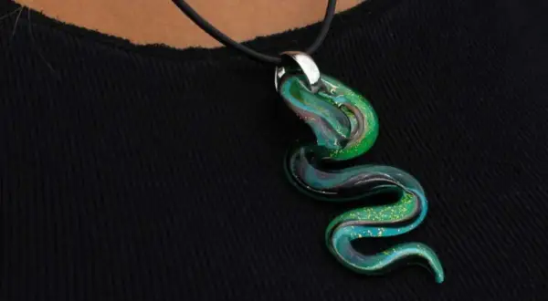 Un serpente sinuoso de **colgante de cristal de Murano** en tonos verdes y azules, con destellos dorados, descansa sobre una tela negra.