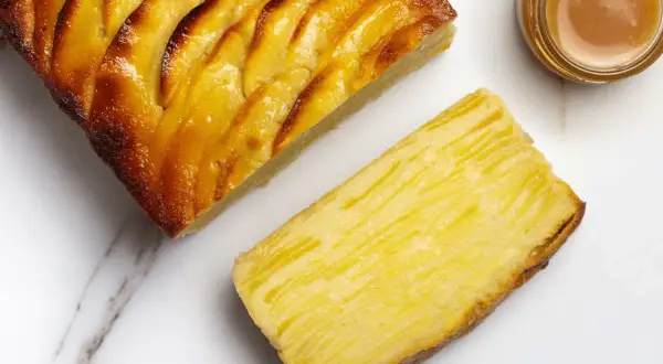 Une tranche de tarte aux pommes, dorée et caramélisée, à côté d'une autre tranche montrant la texture fine des pommes. Un petit pot de caramel est aussi visible.