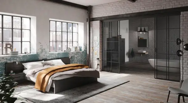 Ein gemütliches **Bett unter Fenster** mit industriellem Charme. Daneben ein offener Badbereich mit Wanne.