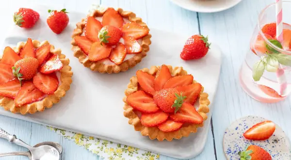 Trois tartelettes aux fraises, garnies de crème et de fruits rouges frais, sur un plateau blanc. Une fraise entière repose à côté.