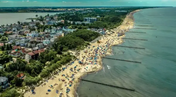 Tłum ludzi na plaży w Mielnie, cieszących się słońcem i morzem. W tle widać zabudowania i jezioro. To idealne miejsce na wakacje, pełne atrakcji.