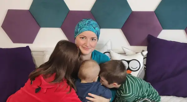 Mama w chustce i trójka dzieci. Najstarszy syn całuje maluszka, córka przytula się do mamy. W tle heksagony i poduszki. BIRADS 4a.