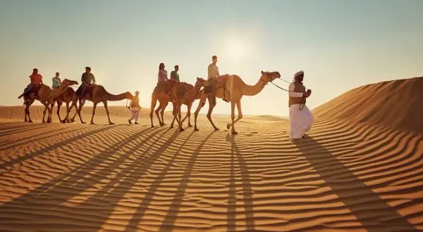 Kamelkarawane in der Wüste bei Sonnenuntergang. Ein Führer in traditioneller Kleidung begleitet Touristen. Tolle Abu Dhabi Tipps für ein unvergessliches Erlebnis.