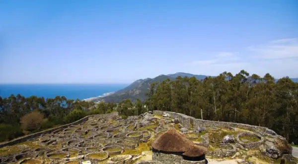 Ruinas circulares de piedra en el monte Santa Tecla, con el mar y montañas al fondo.